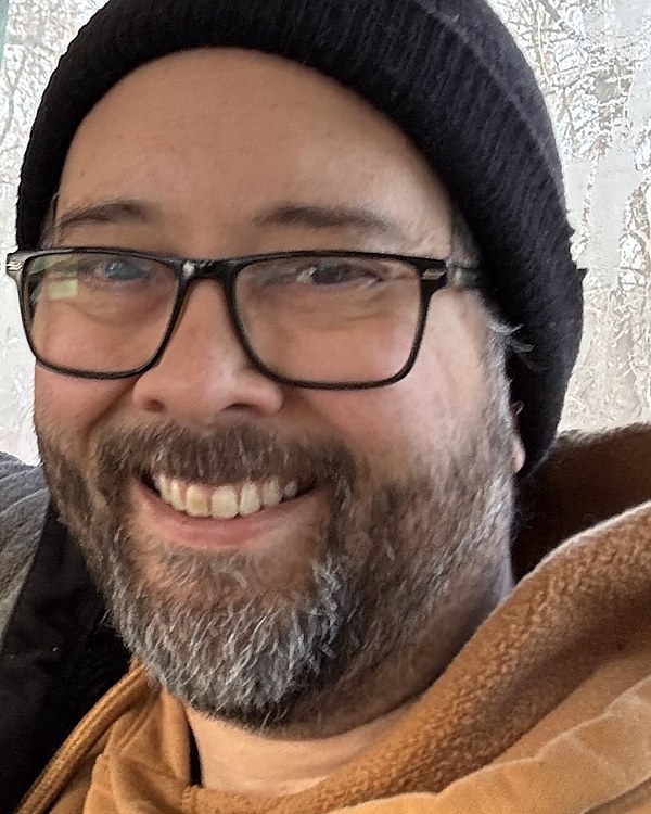 Carl Henshaw, in glasses, a beard and a dark knit hat, smiling aboard an excursion coach in winter.