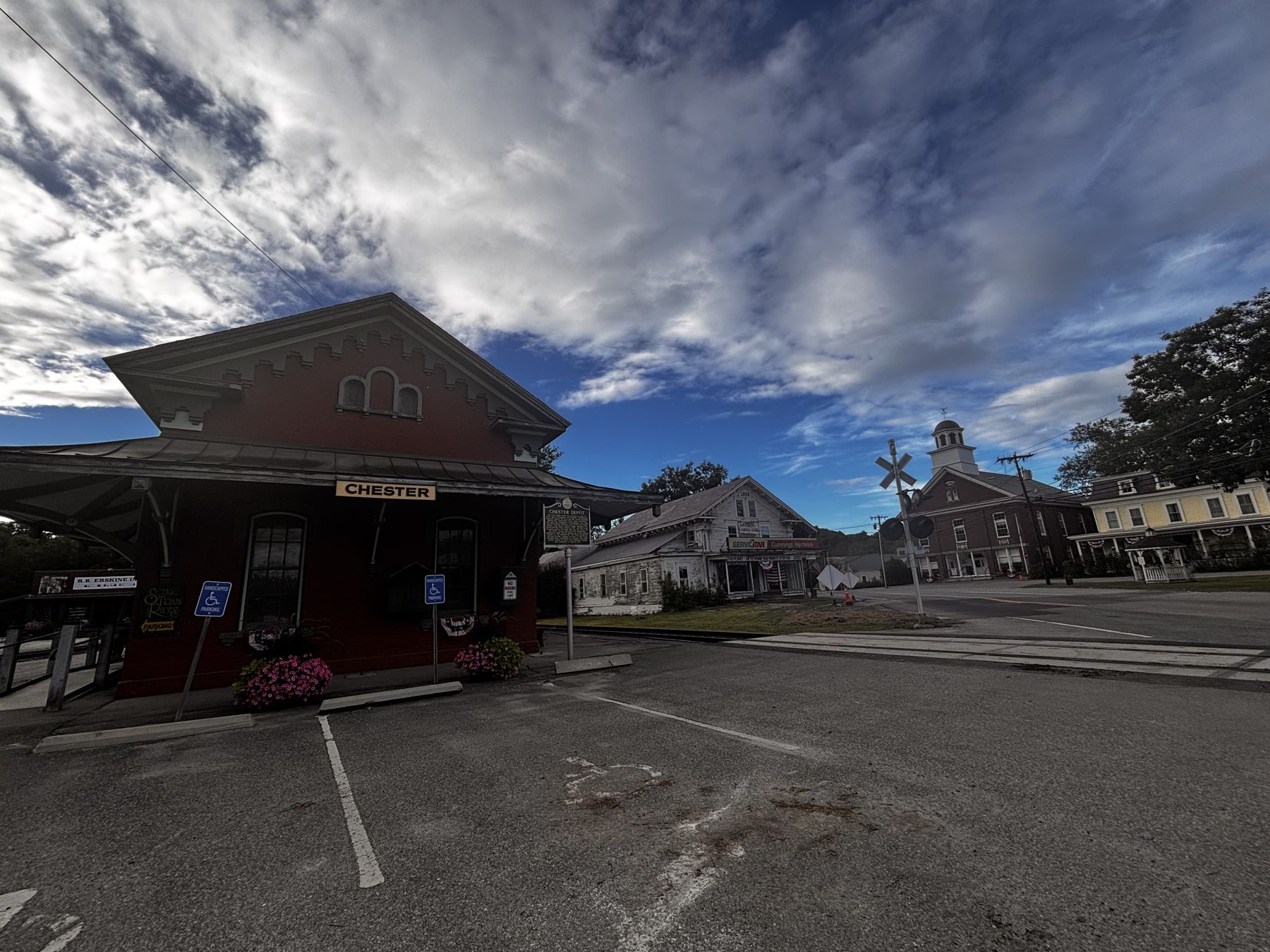 Chester Depot village: the brick station at left, a white hardware store, the grade crossing, and the brick town building with its white cupola under a wide sky.