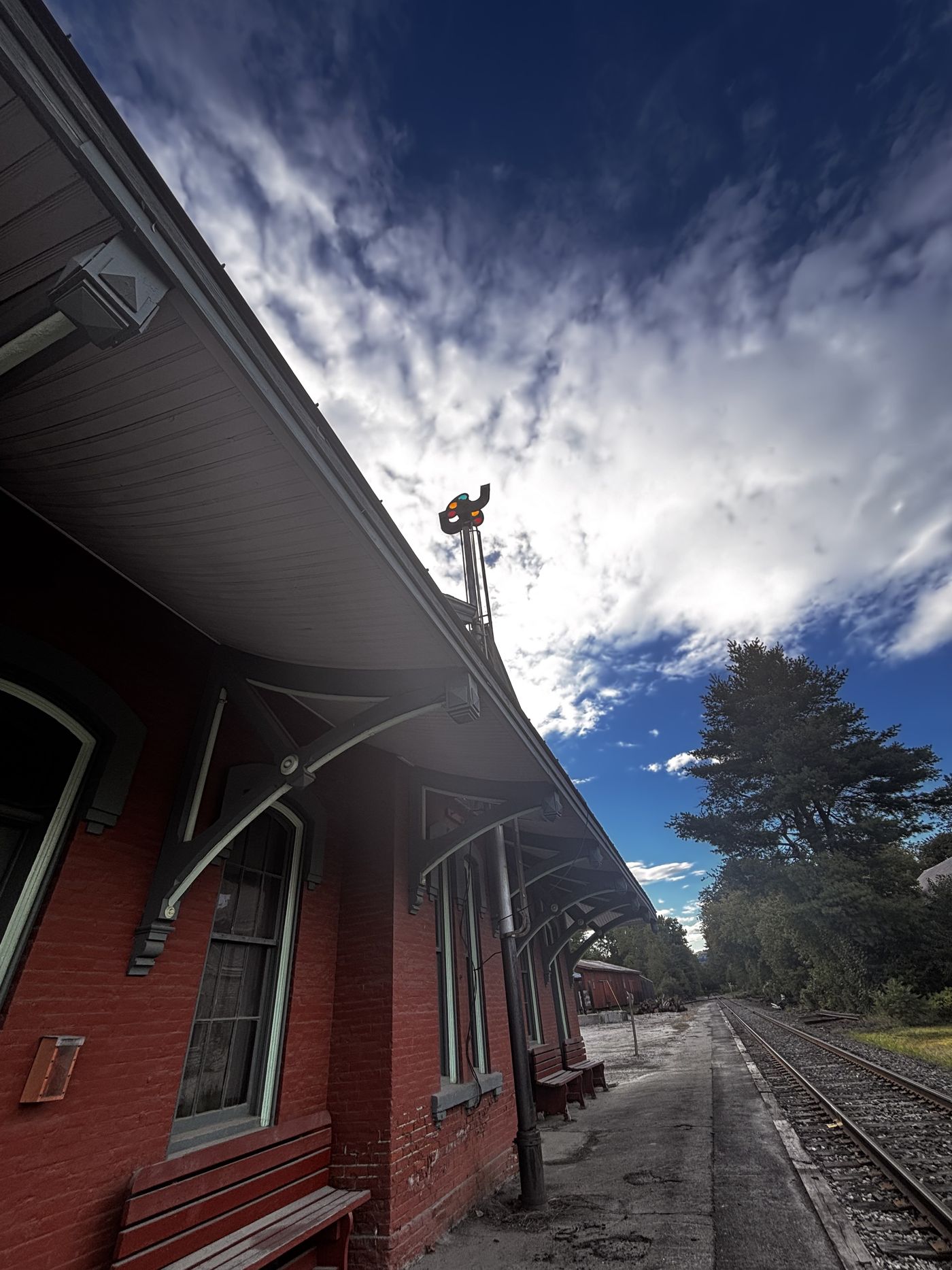 Looking up along the eaves of Chester Depot toward a color-light signal, the track curving away beside the platform under a dramatic sky.