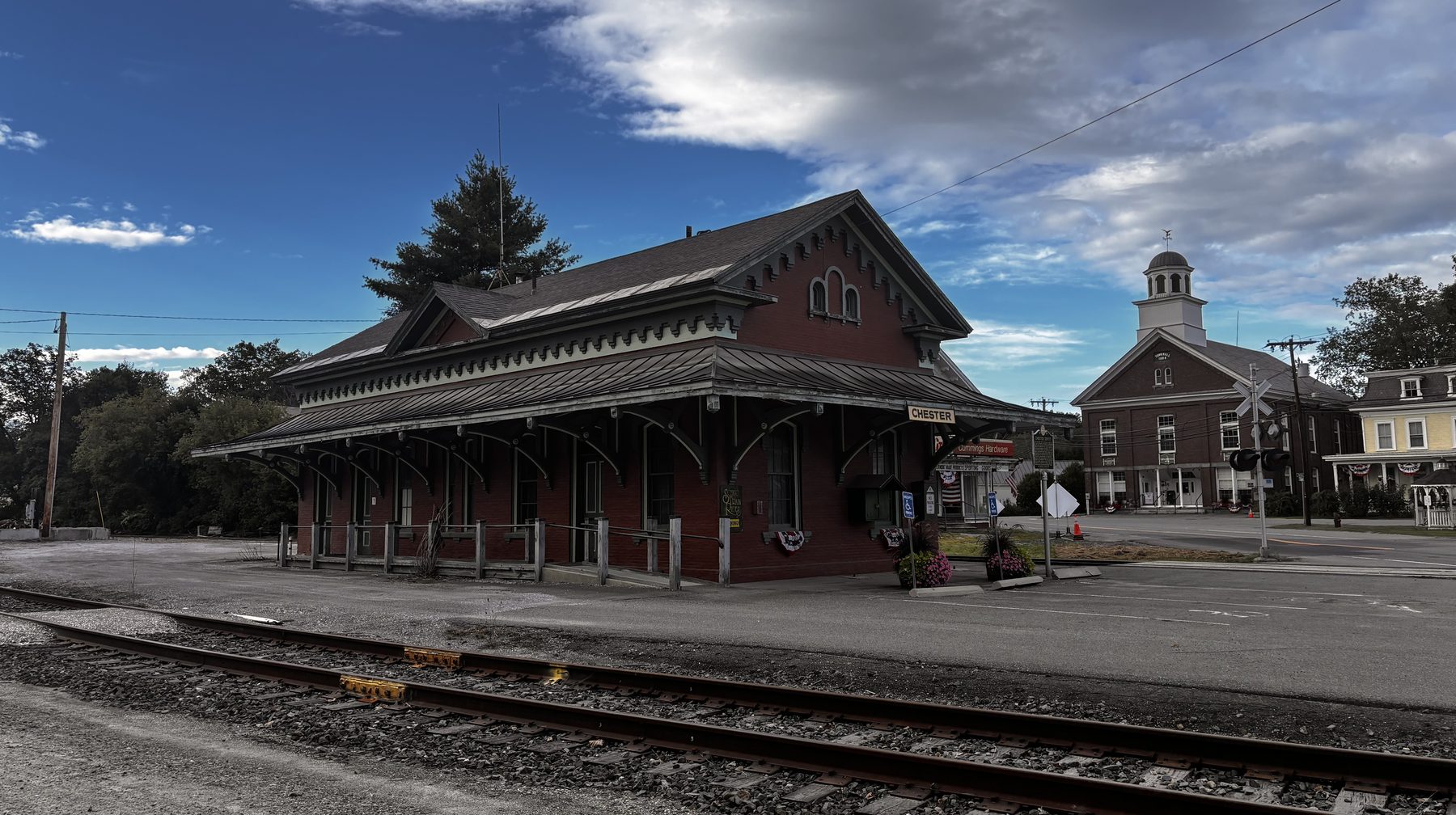 Chester Depot, an 1871 brick Italianate station with a wide bracketed roof, seen across the tracks with the village cupola behind it under a bright, clouded sky.